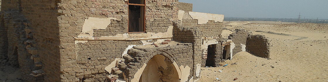 View of partially preserved mudbrick and stone structures at the archaeological site, illustrating the effects of time, sand accumulation, and ongoing conservation efforts.